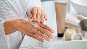 Adult hands applying rich unbranded hand cream beside a balm jar and towel on a bright bathroom counter