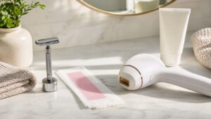 Unbranded razor, wax strip, IPL-style device, towel, and moisturizer on a bright bathroom vanity